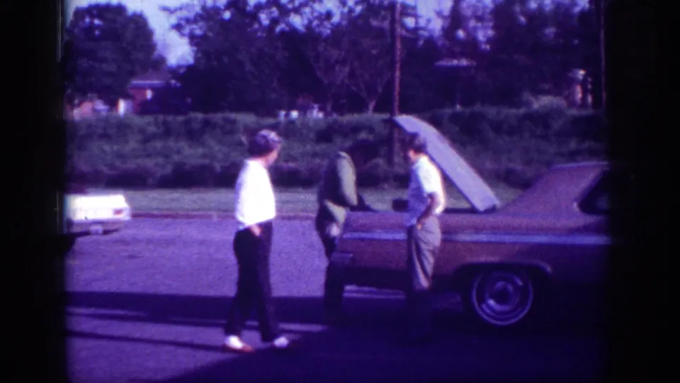 Three men gathered around an open-hood early-1960s American sedan on a residential street in Memphis, Tennessee, 1963, captured in aged 8mm film with a strong purple-blue color cast.