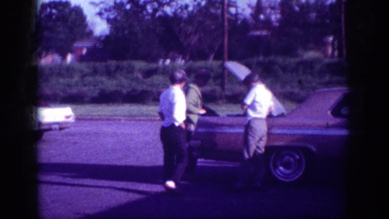 Two men standing beside an open-hood 1960s American sedan; a third man partially visible at right.