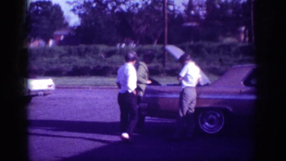 Similar grouping of three men around an open-hood early-1960s sedan on a suburban Memphis street, with slight camera repositioning visible in this aged 8mm home-movie frame.