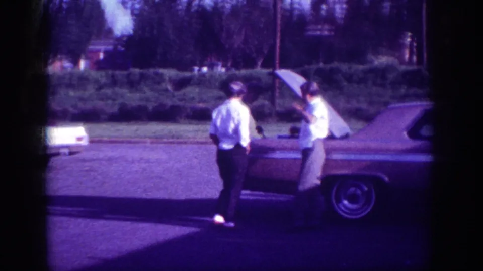 Two men visibly engaging with the open engine area of a large early-1960s American sedan, filmed in aged 8mm on a Memphis residential street circa 1963.