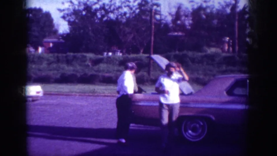 Two men bending toward the open engine bay of a large early-1960s American sedan in a suburban Memphis street scene, filmed on aged 8mm with a characteristic violet color cast.