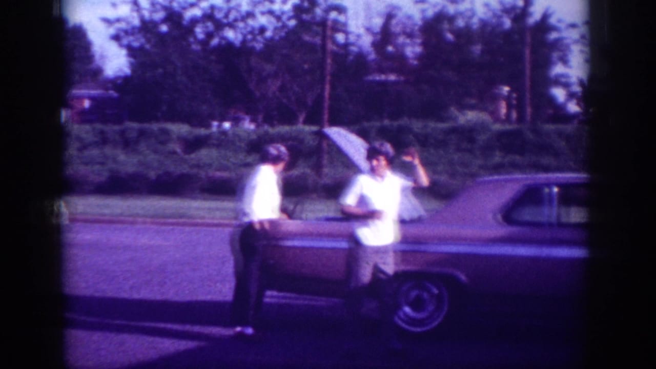 Two adult men beside what appears to be an early-1960s American hardtop sedan on a suburban street in Memphis, Tennessee, captured on aged 8mm film with a pronounced purple color cast.