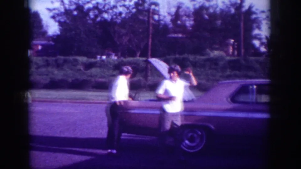 Two adult men beside what appears to be an early-1960s American hardtop sedan on a suburban street in Memphis, Tennessee, captured on aged 8mm film with a pronounced purple color cast.