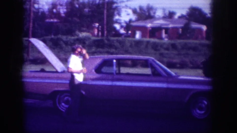 A man in a white shirt stands at the open hood of a large early-1960s-style American sedan on a suburban street in Memphis, Tennessee, shot on 8mm film with a strong purple-violet tint.