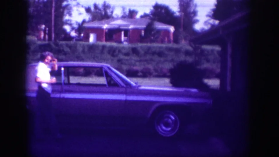 A man leans against the rear section of a large two-door American sedan parked on a residential street in Memphis, Tennessee, with a brick house and clipped hedges behind; footage shows aged 8mm purple hue.