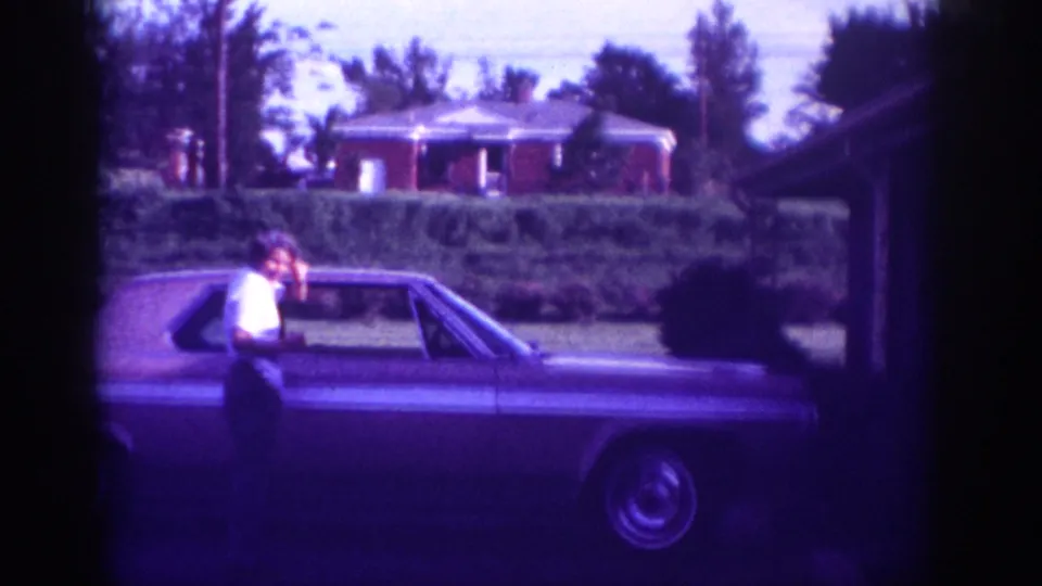 A man sits or leans on the rear of a large American sedan on a residential street, identified in metadata as Memphis, Tennessee, ca. 1963; pronounced purple-violet 8mm color cast throughout.