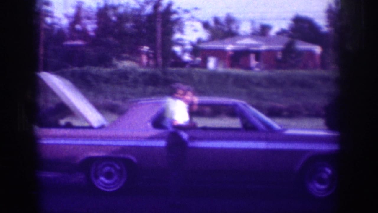 Man standing beside a long car with its hood open on a residential street.