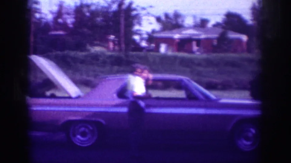 In supplied 1963 Memphis home-movie footage, a man in a light shirt stands beside a long two-door car with the hood raised on a suburban street.