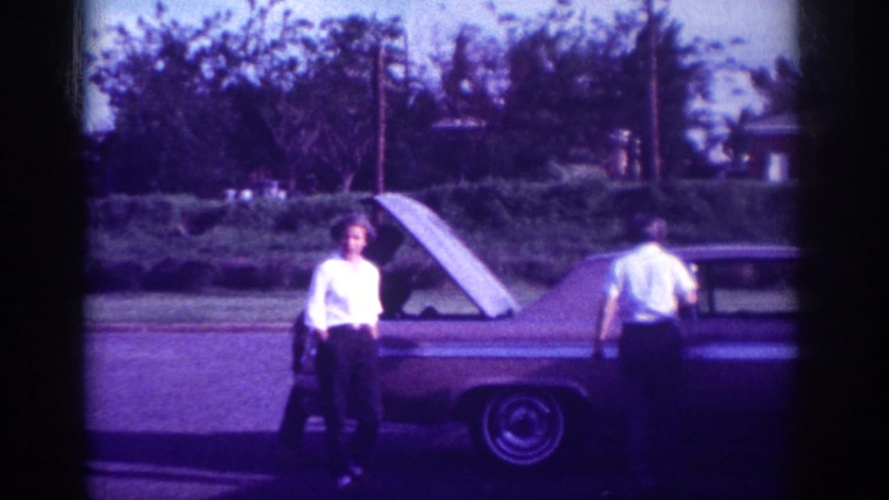 Two adults stand near a car with its hood open on a quiet street.