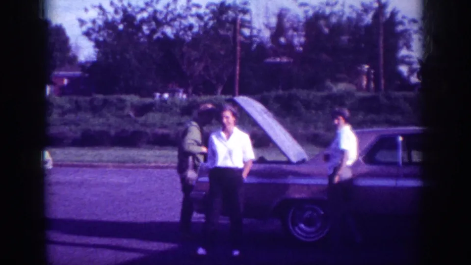 Three adults appear around the open hood of a stopped car on a residential street, with one person facing the camera and another near the driver's side.