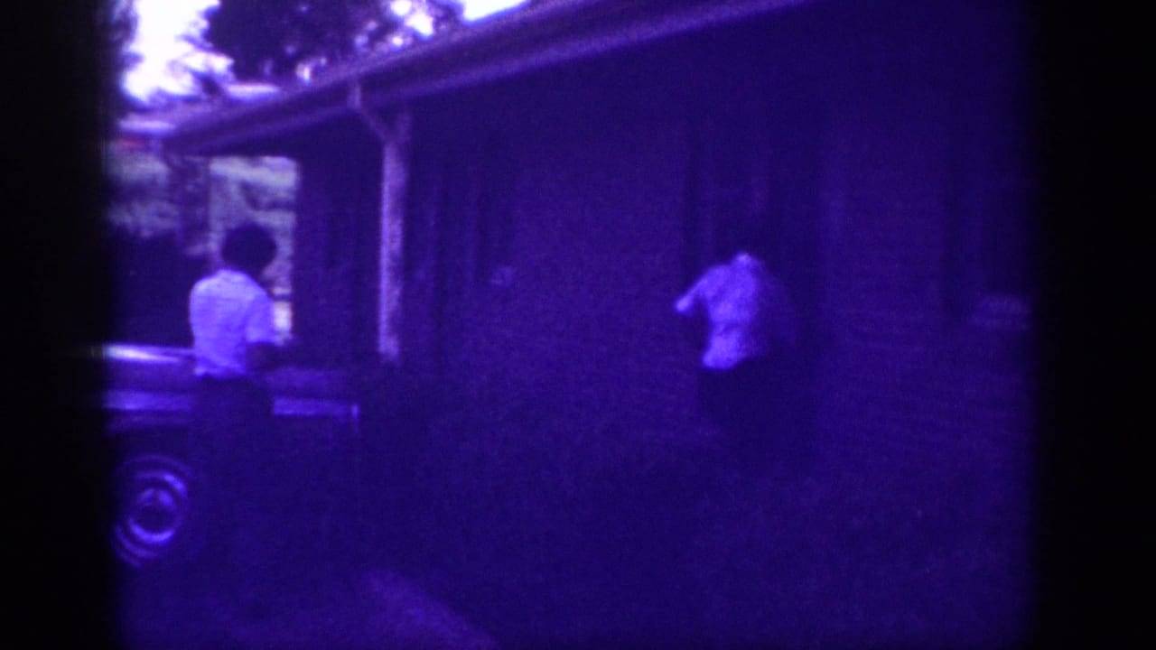 Two figures in white shirts approach a ranch house entrance; partial car wheel visible at lower left; heavy purple dye-fade cast.