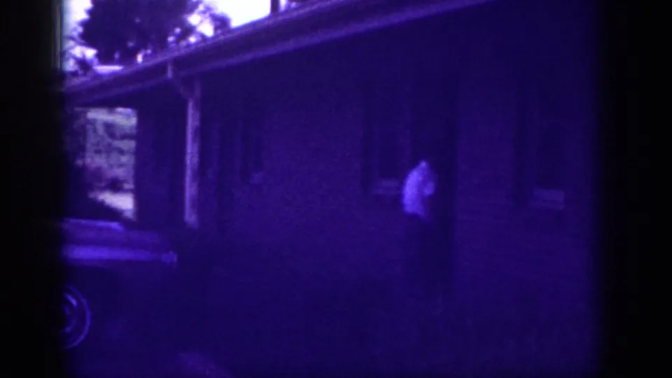 Figure in light clothing near a house entrance with car partially visible at lower frame, Memphis Tennessee circa 1963; dark purple color cast from film aging.