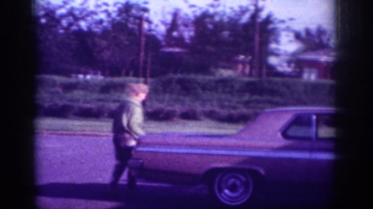 Woman in light jacket approaching trunk of a large early-1960s American sedan on a Memphis residential street, ca. 1963; 8mm color film with violet color shift.
