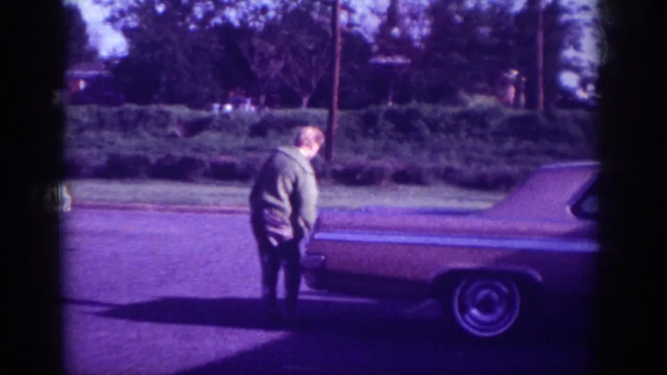 Woman standing at rear of early-1960s American sedan on residential street, Memphis, ca. 1963; hedgerow and utility poles in background; 8mm film with violet color cast.