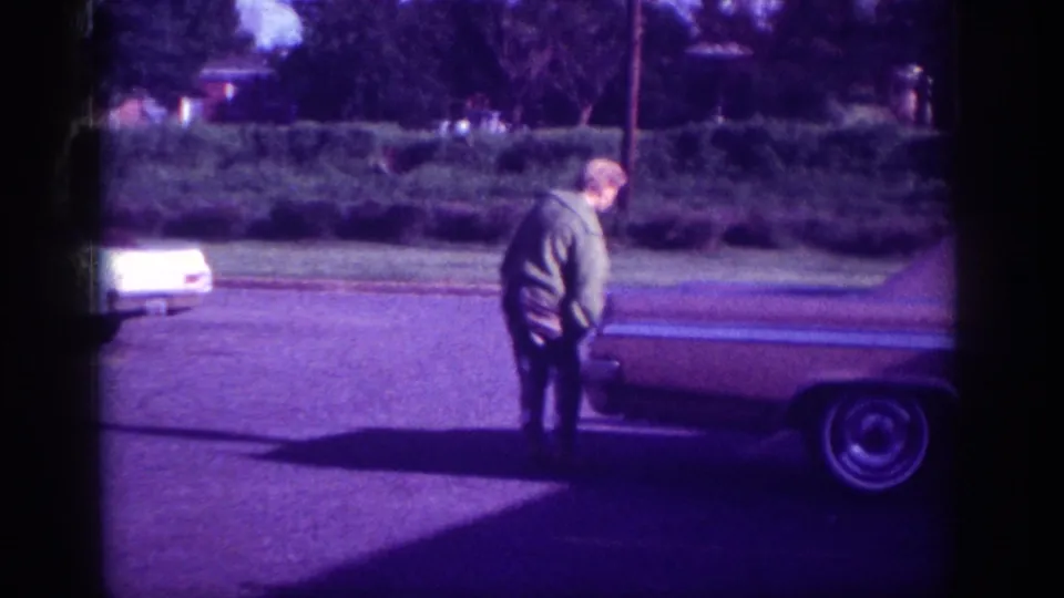 Woman at trunk of early-1960s sedan, Memphis, ca. 1963; a second white vehicle is visible in the background; 8mm footage with aged violet color shift.