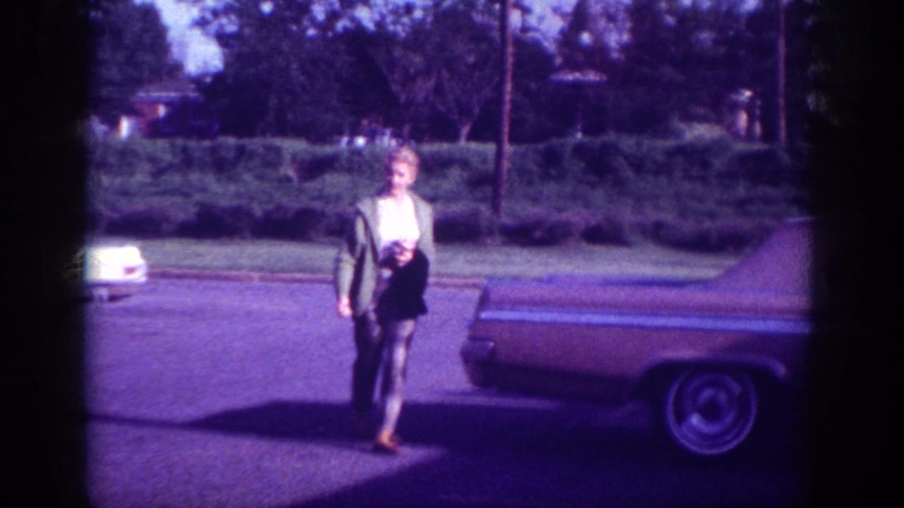 Woman in dark jacket standing upright near rear of American sedan, turning toward camera.