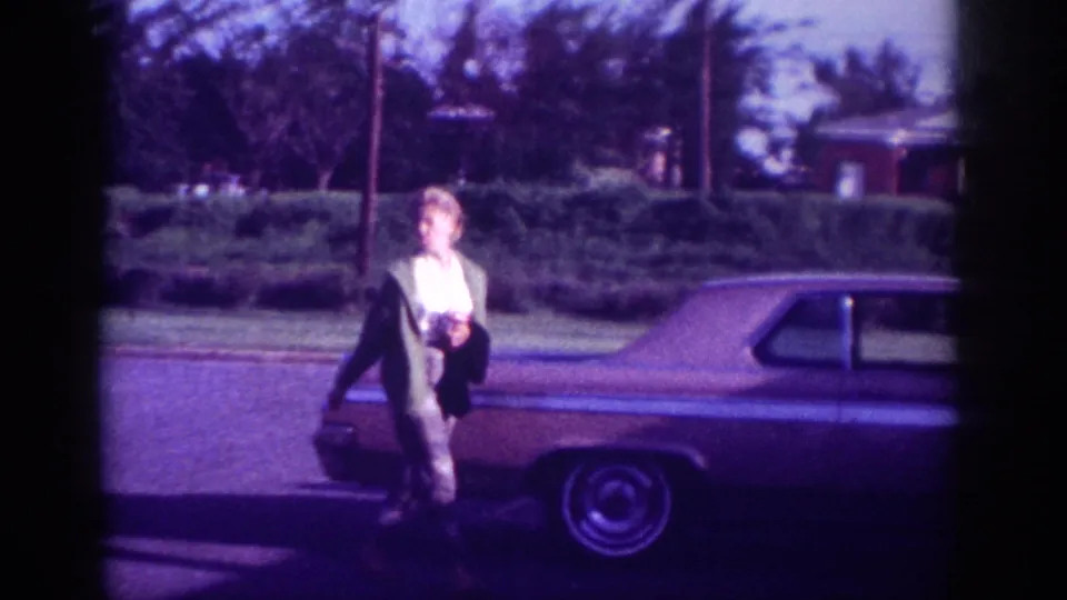 Woman in dark jacket walking along the side of a large early-1960s American sedan, trees and brick structure visible, suburban Memphis setting circa 1963.