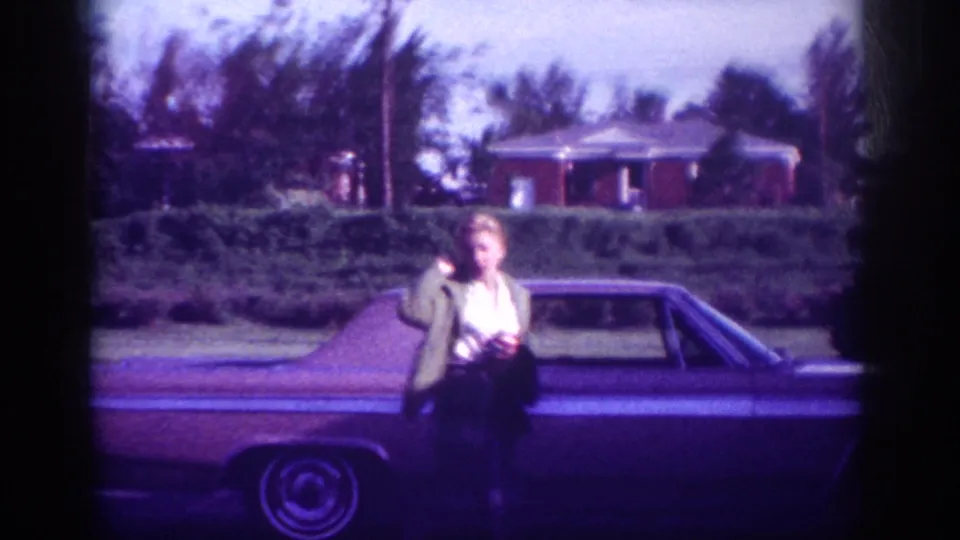 Woman in dark jacket standing beside a large early-1960s American sedan facing camera, brick house and mature trees behind her, suburban Memphis circa 1963.