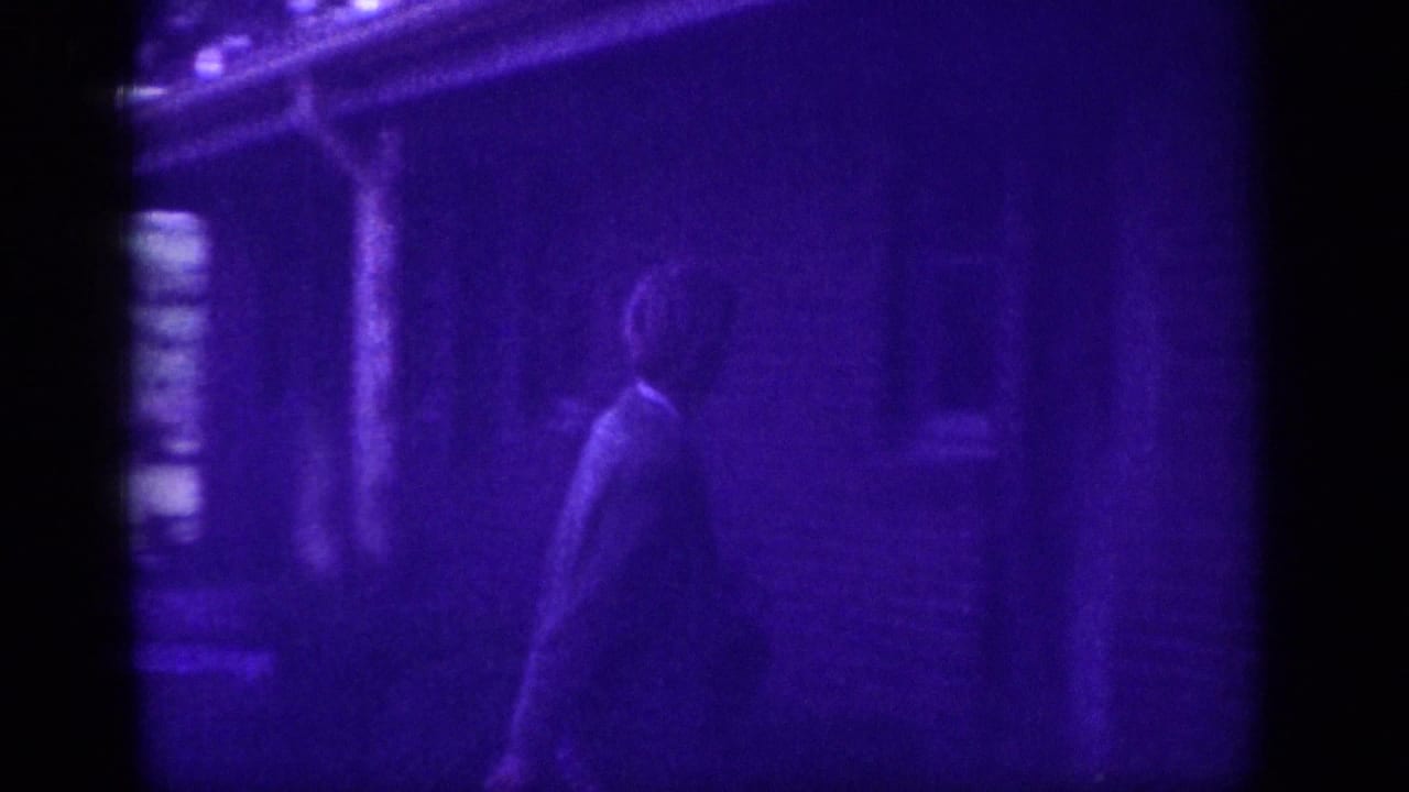 Dimly lit figure walking near a structure, heavy blue-violet color cast from aged 8mm film.