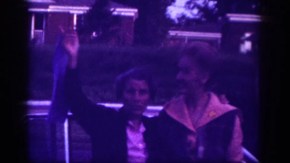 Two women stand on a suburban lawn in this 1963 8mm home movie; a single-story house with a lit porch and mature trees is visible behind them.