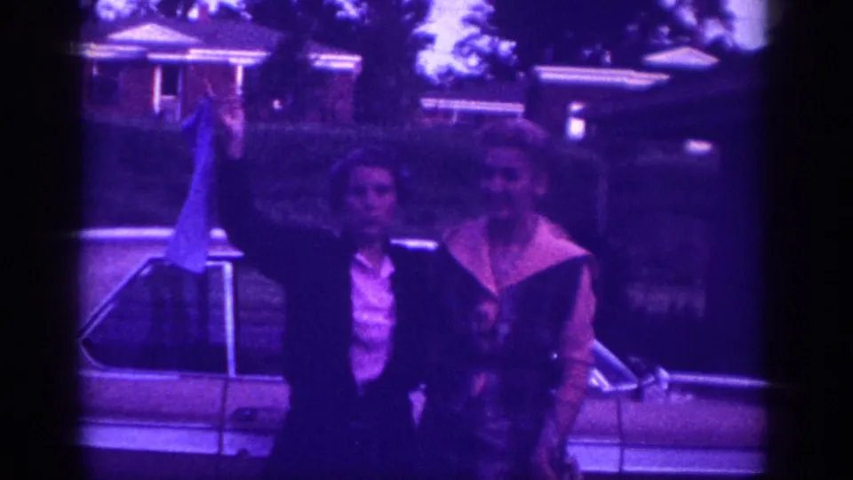Two women pose in front of a light-colored classic American car in this 1963 Memphis home movie; one lifts a blue fabric piece while both face the camera.