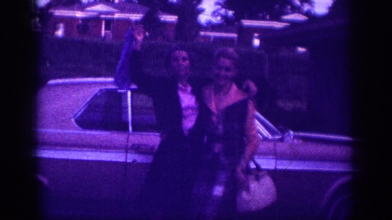 Two women in early-1960s dress pose beside a large American sedan in a suburban Memphis, Tennessee neighborhood, c.1963; heavy purple dye shift from aged 8mm stock.
