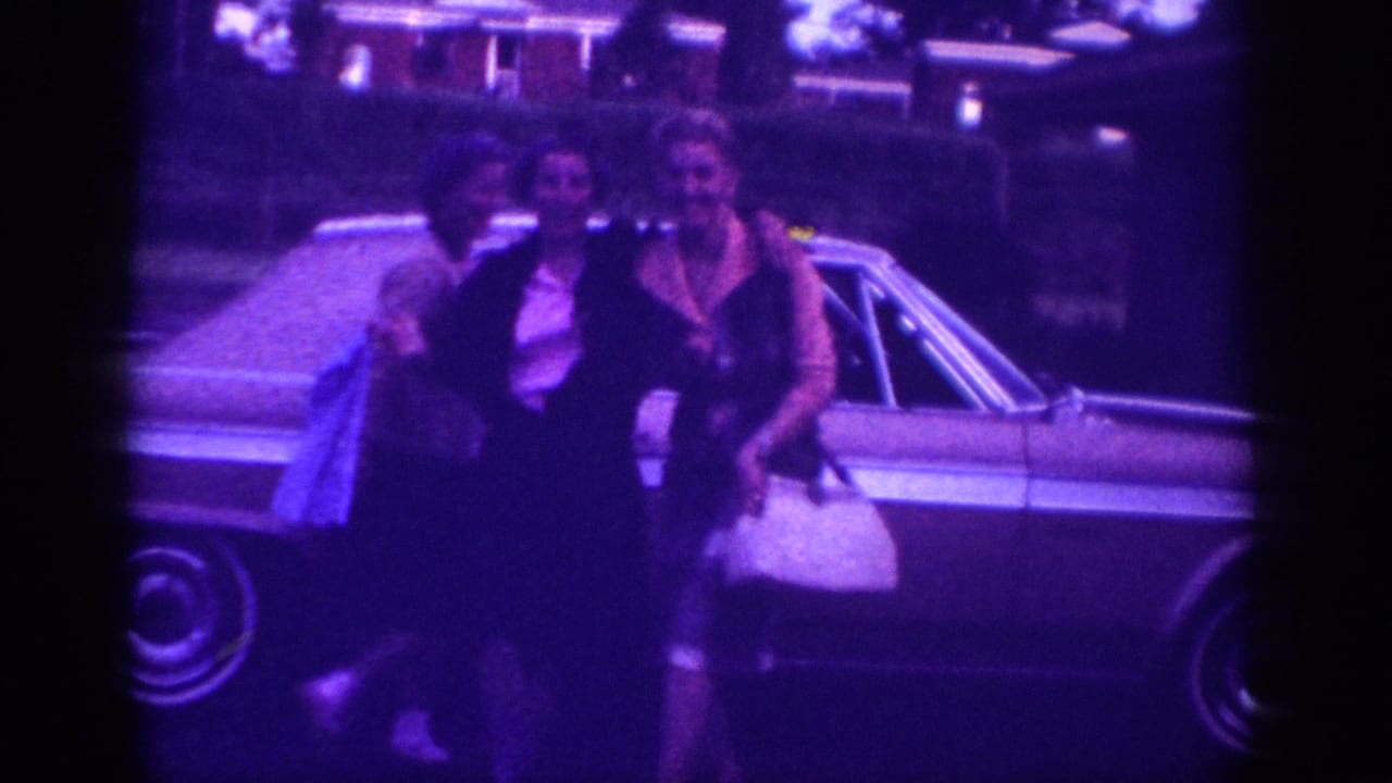 Two women in 1960s attire pose near a large American sedan in Memphis, Tennessee, c.1963; one carries a white handbag; purple film cast from aged 8mm stock.
