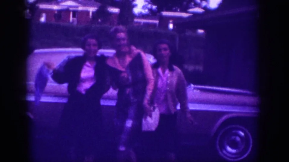 Three women in early-1960s dress stand before a large American sedan in suburban Memphis, Tennessee, c.1963; one raises her arm toward the camera.