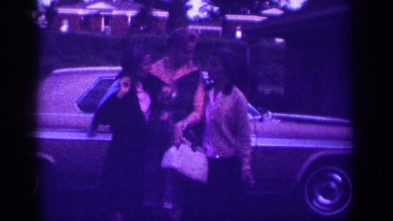 Three women in early-1960s blouses and jackets pose beside a full-size American sedan in what may be a Memphis, Tennessee suburban street, circa 1963; heavy violet cast from aged 8mm color stock.