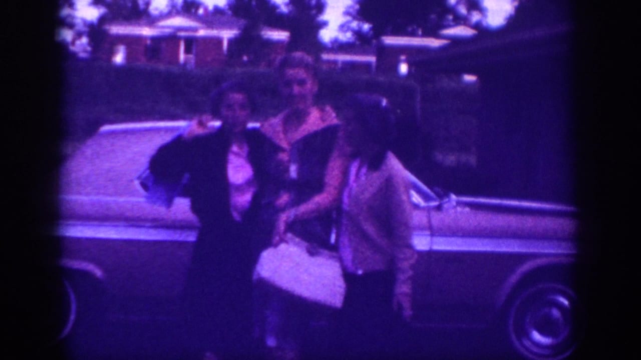 Three women in early-1960s dress pose beside a full-size American car in a suburban setting, circa 1963; one woman holds a white handbag; characteristic violet dye-shift from aged 8mm stock.