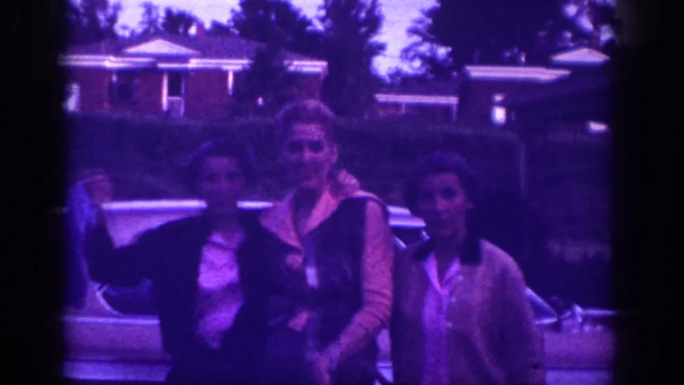 Two women in 1960s clothing smile toward the camera in front of a large American sedan and brick suburban houses, possibly Memphis, Tennessee, circa 1963.