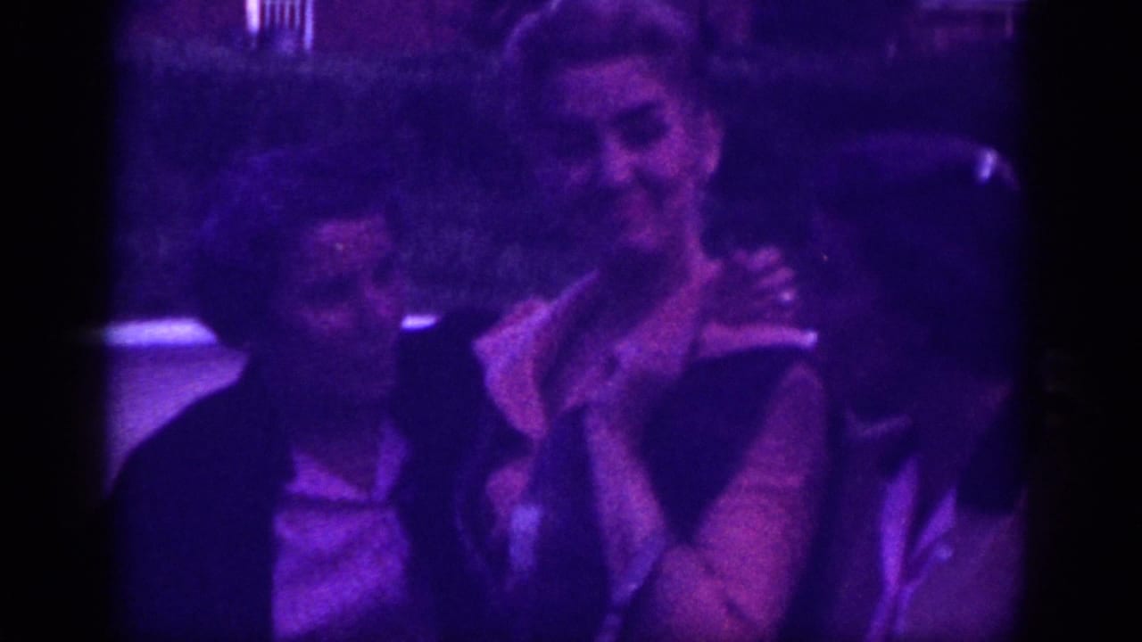 Two women laugh together in a tighter shot, brick building visible behind, on purple-shifted 8mm film.