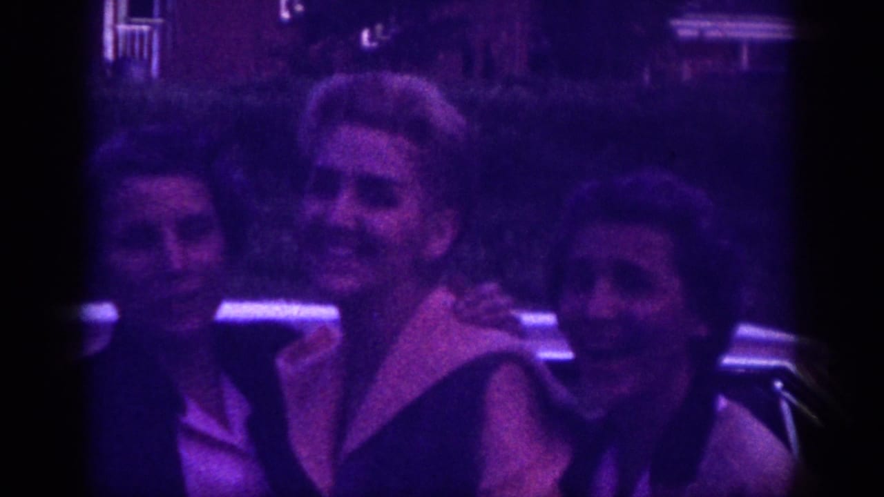Three adult women sharing a joyful moment beside a car in an evening setting, shot on 8mm in Memphis, Tennessee, circa 1963, with characteristic purple film dye-fade.