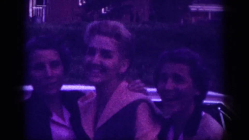 Three adult women sharing a joyful moment beside a car in an evening setting, shot on 8mm in Memphis, Tennessee, circa 1963, with characteristic purple film dye-fade.