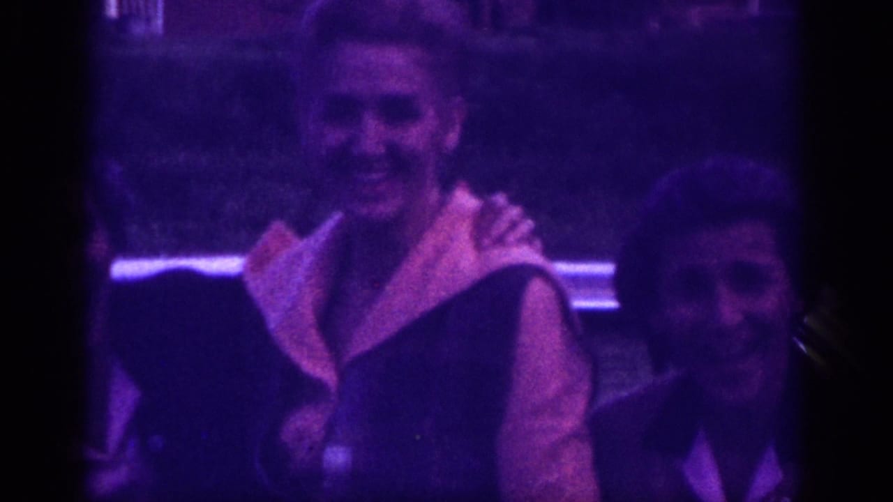 Two women in close embrace, one in dark dress with light lapel panel, nighttime shot with heavy purple cast on 8mm film.