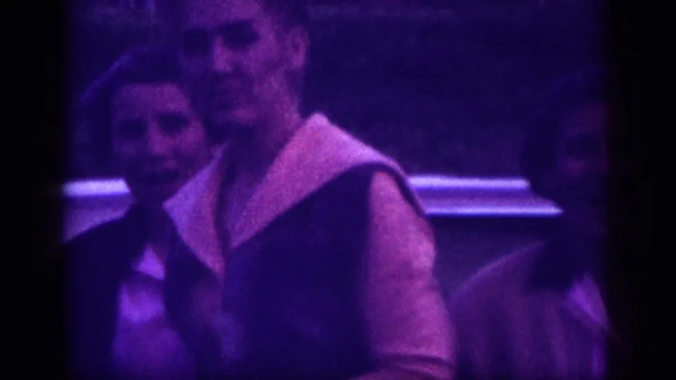 Close framing on a smiling adult woman in 1960s dress with a distinctive light collar, second figure partially in frame; 8mm nighttime footage with strong purple color shift, circa 1963.