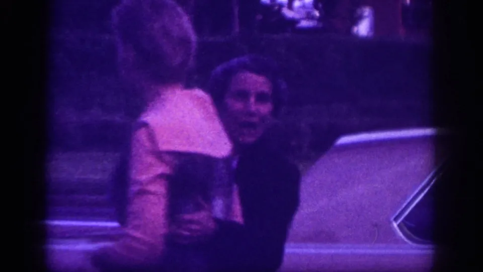 Two smiling adult women standing beside a period car in an evening setting, likely Memphis, Tennessee, circa 1963; car chrome or body panel visible at right frame edge; 8mm stock with purple dye-fade.
