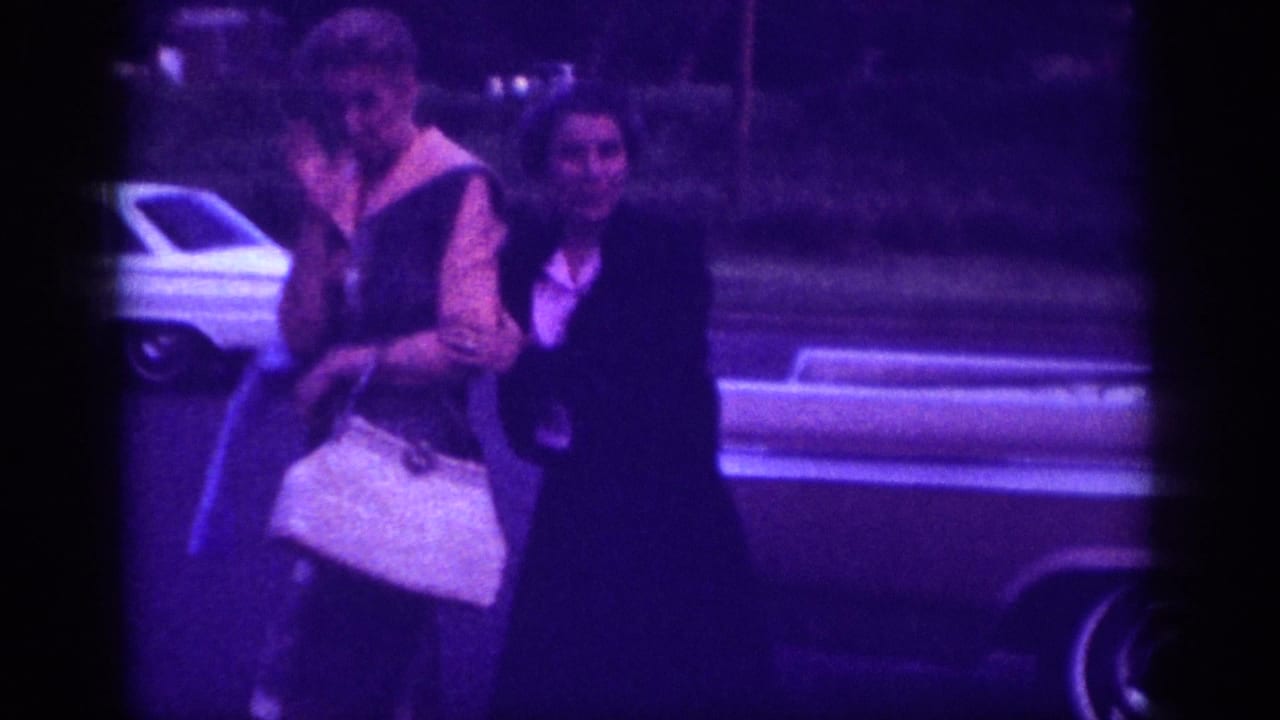 Two women walk alongside a white American sedan with chrome trim, one carrying a structured white handbag; Memphis, Tennessee, circa 1963, 8mm home movie.