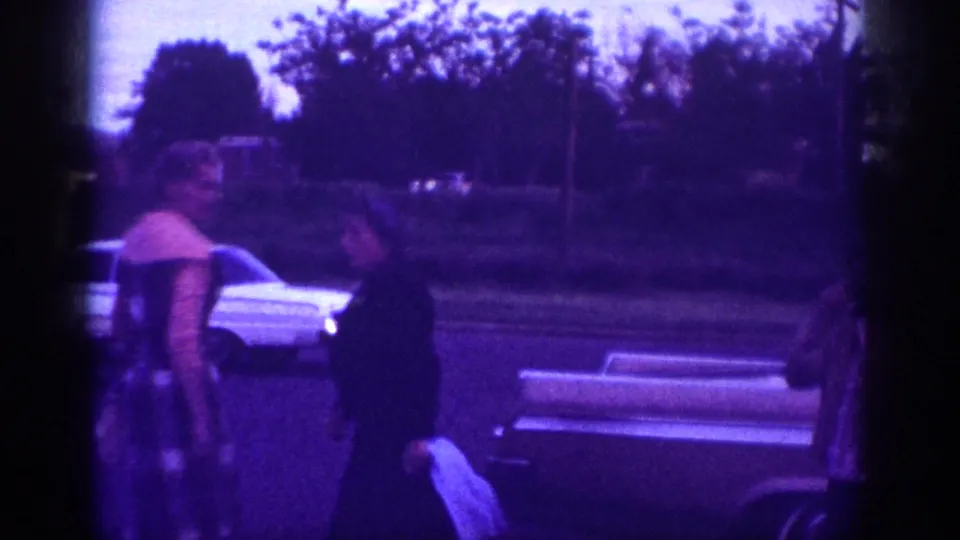 A woman walks along a street near parked cars and foliage at dusk; Memphis, Tennessee area, circa 1963, 8mm home movie.