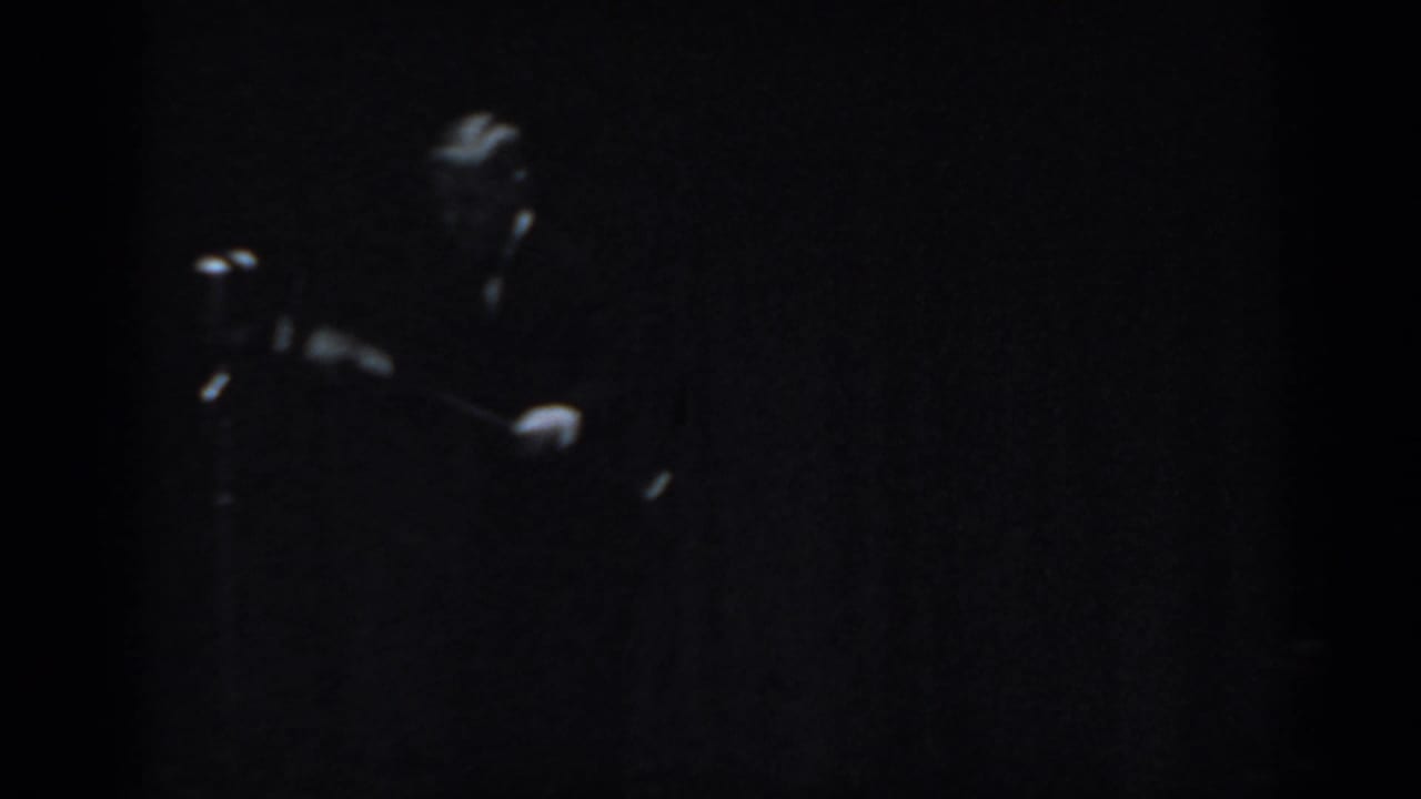 Near-silhouette of a male figure in dark clothing against a black background, captured in very low light with heavy 8mm grain.
