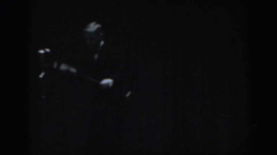 Male performer in dark clothing shot as near-silhouette under very low stage lighting on 8mm film, consistent with 1965 Saint Louis live entertainment.