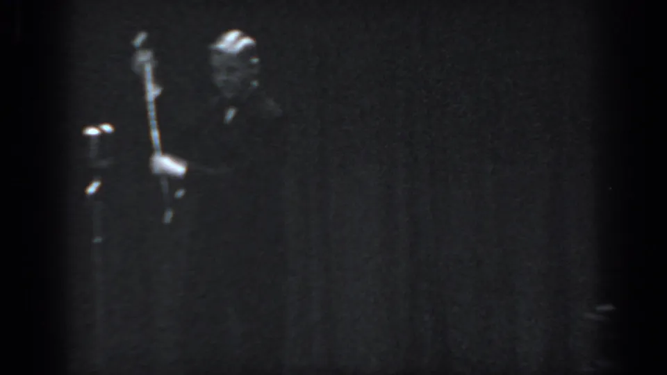 Male performer in dark suit beside a microphone stand on a dimly lit stage, captured on 8mm in Saint Louis, 1965, with a vertical-draped curtain backdrop visible.