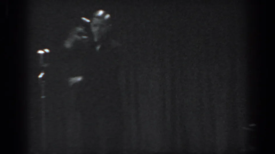 Male performer in motion on a dimly lit stage beside a microphone stand, 8mm footage consistent with 1965 Saint Louis variety entertainment.