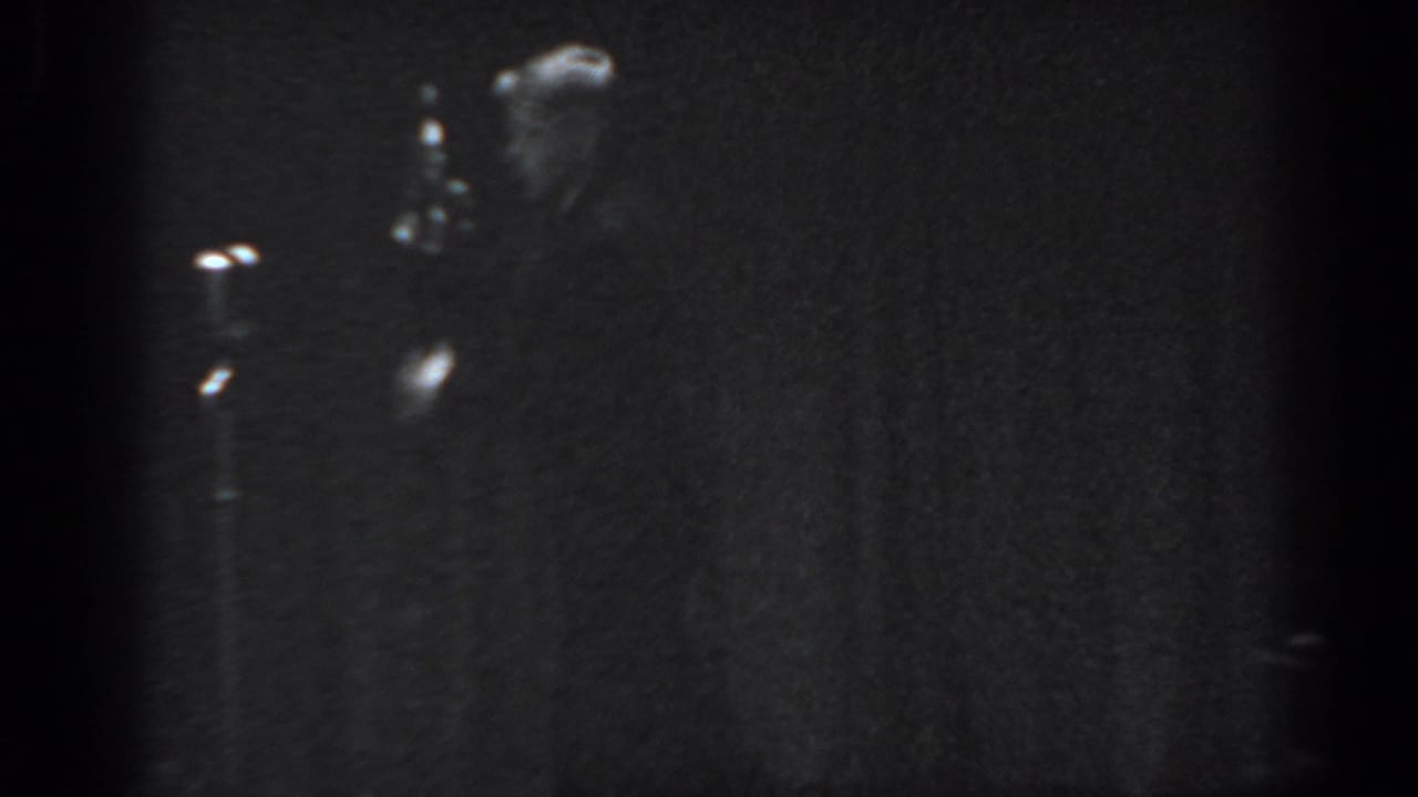 Heavily underexposed 8mm frame from what appears to be a 1965 Saint Louis stage performance, showing a silhouetted male performer against a dark draped backdrop.