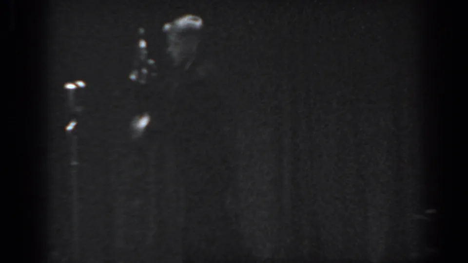 Heavily underexposed 8mm frame from what appears to be a 1965 Saint Louis stage performance, showing a silhouetted male performer against a dark draped backdrop.