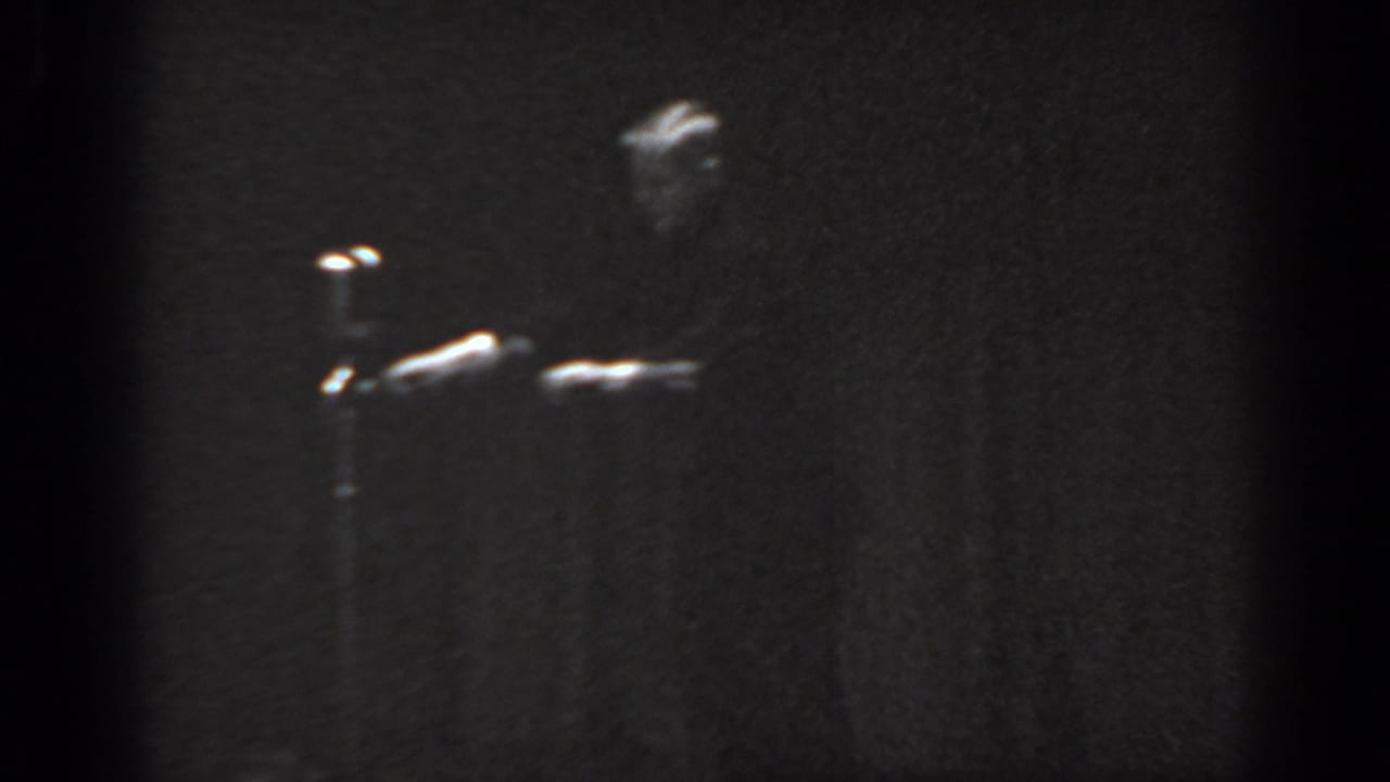 8mm film frame circa 1965 showing a male performer near stage right with a microphone stand visible, heavy grain, and underexposure from indoor amateur filming.