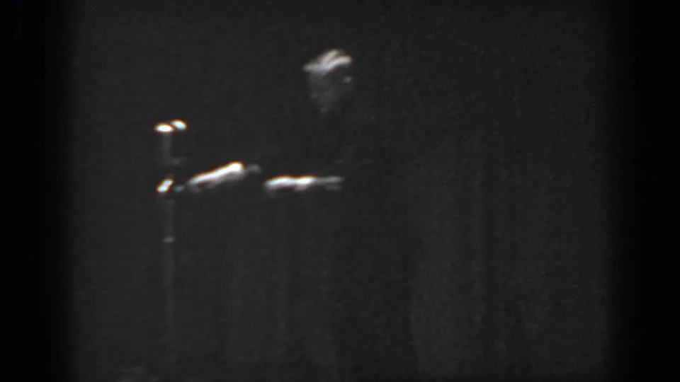 8mm film frame circa 1965 showing a male performer near stage right with a microphone stand visible, heavy grain, and underexposure from indoor amateur filming.