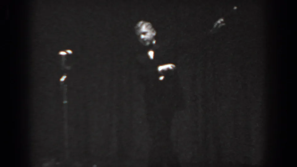8mm frame from a 1965 Saint Louis stage performance showing a male performer's face partially illuminated, a microphone stand at left, and a second figure partially visible.