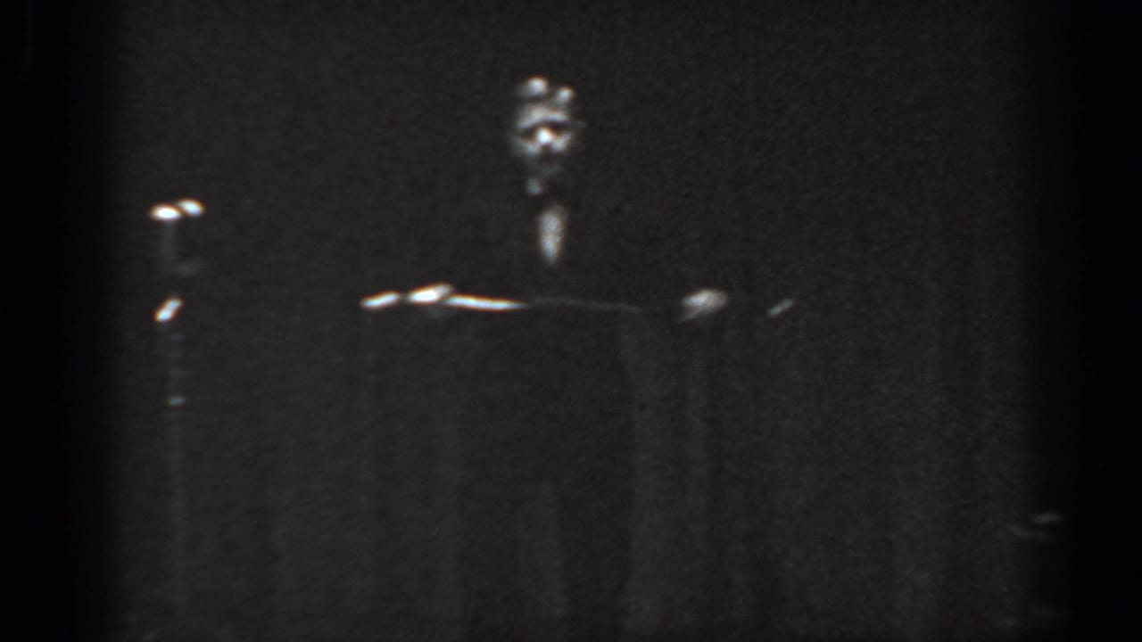 Dark 8mm stage frame with a distant figure near a raised object, bright stage-light glints, and a dark curtain backdrop.