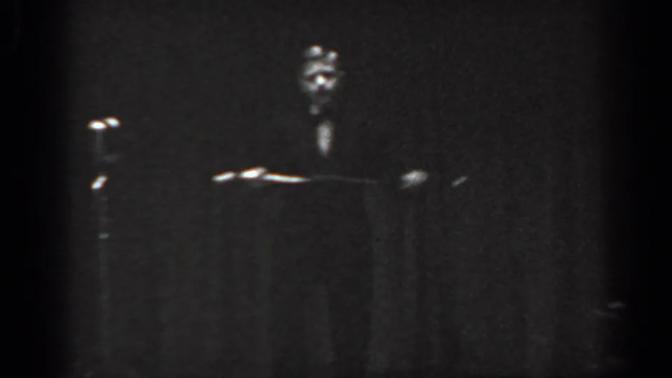 Black-and-white 8mm home movie frame, circa 1965, of a dimly lit stage with a small figure near a podium-height object and stage lights glinting in the background.